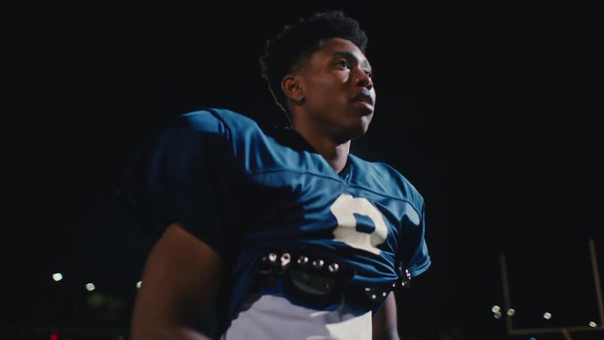 A teenage boy in a blue football jersey throwing a football to someone on a field at night