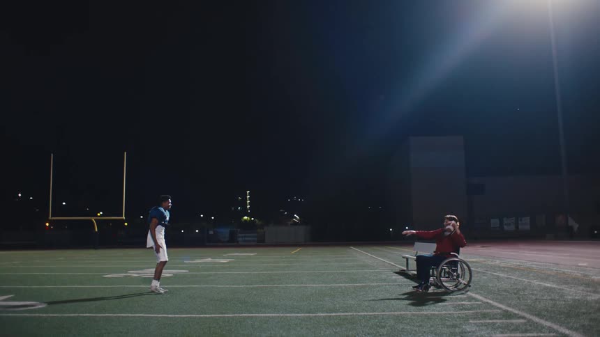 Person in wheelchair throwing an american football to a uniformed man on a field at night
