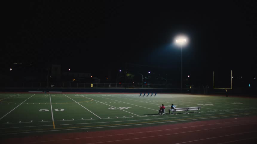 A nighttime scene at an outdoor american football field illuminated by bright lights