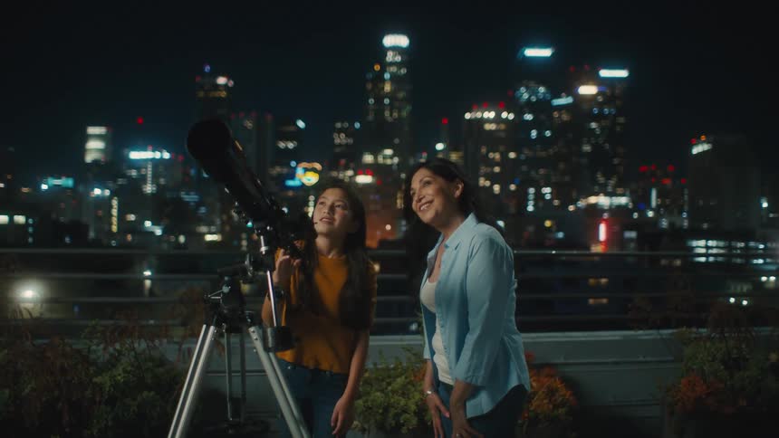 Two women are looking at a telescope pointed at the sky one night and behind them are several large buildings with lights on