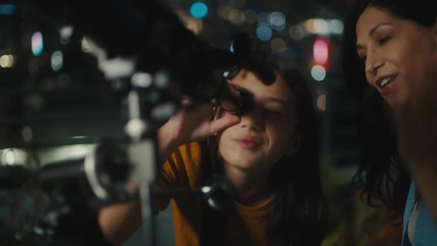 A young girl and woman look through a telescope at night with the city lights in the background