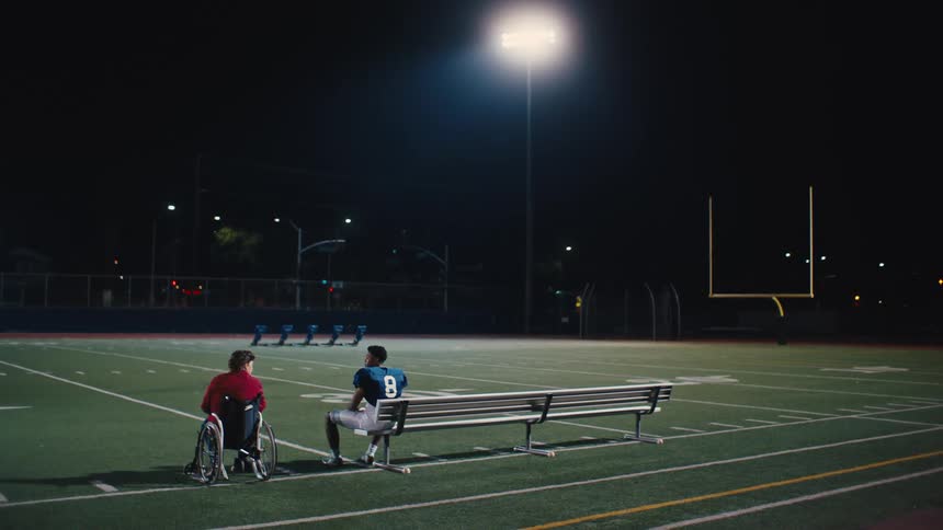 Uniformed man sitting on a bench on an american football field and next to him a person in a wheelchair
