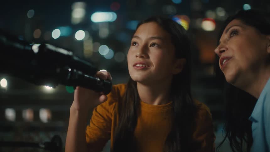 A young female looks through a telescope while outdoors with an older woman