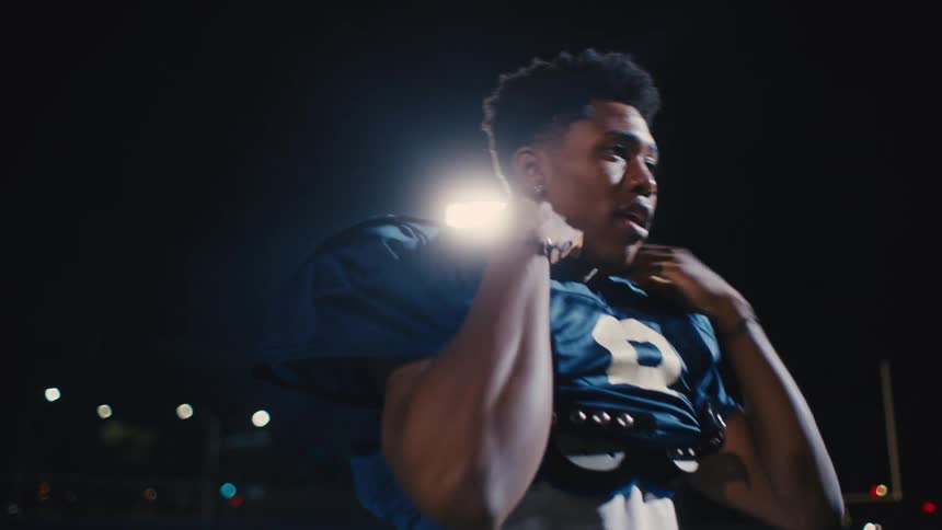 A young man in a dark blue sports jersey throwing an american football at night