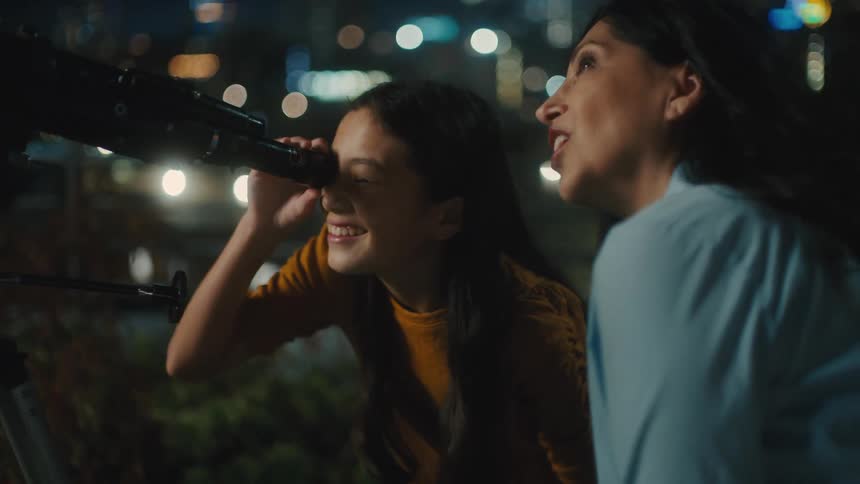 Woman next to a girl looking through a telescope at night