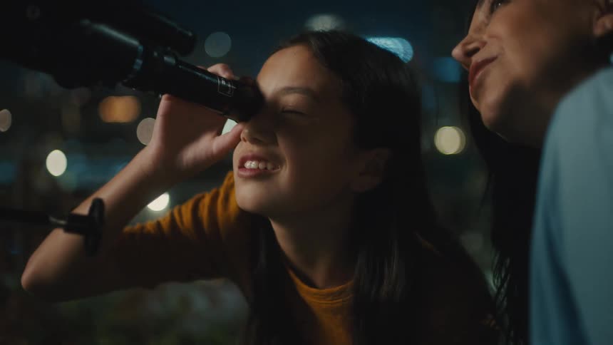 Mother and daughter are looking through a telescope at night in the city