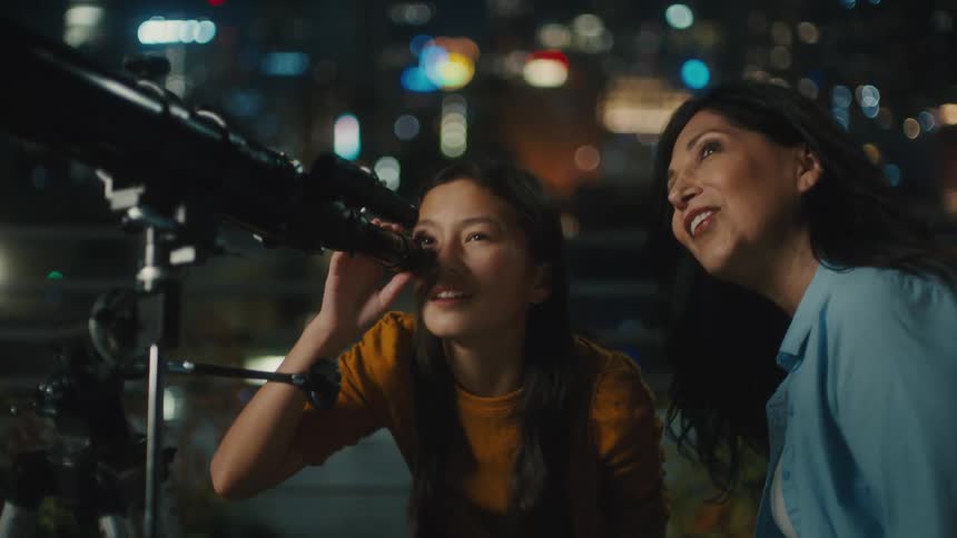 In a city terrace a teenage girl looks into a telescope while her mother talks to her about it and they both smile