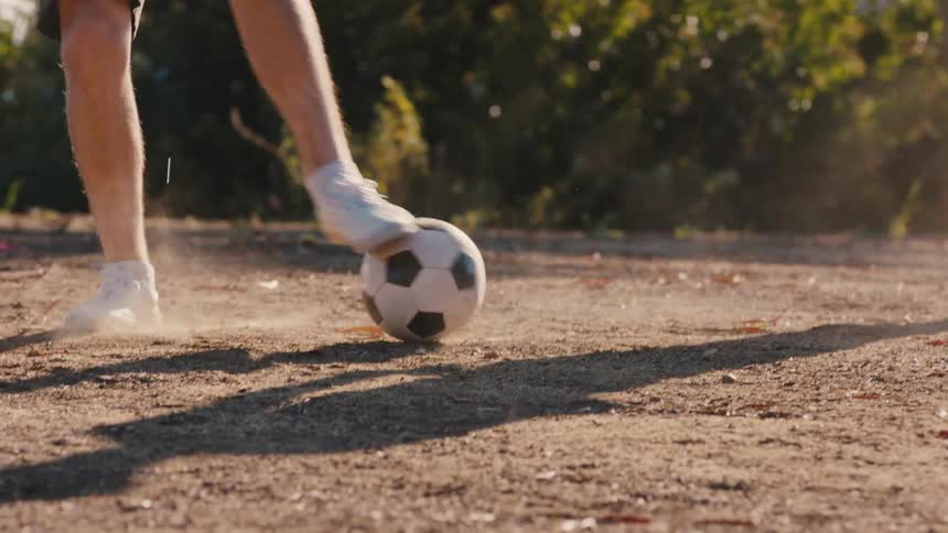Two men kicking a soccer ball around on a gravel area in the middle of the day