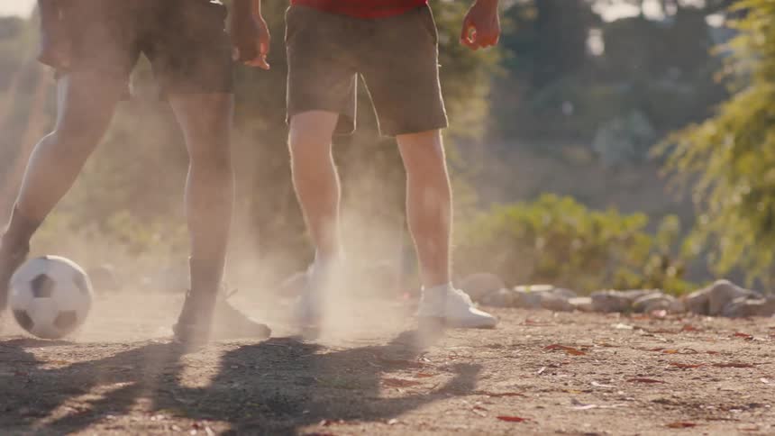 Legs of two different boys playing soccer between each other on a dirt area