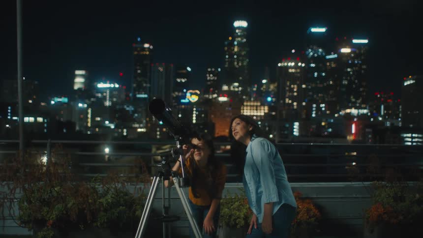 A woman looking through a camera lens while another woman stands next to her with a city skyline in the background