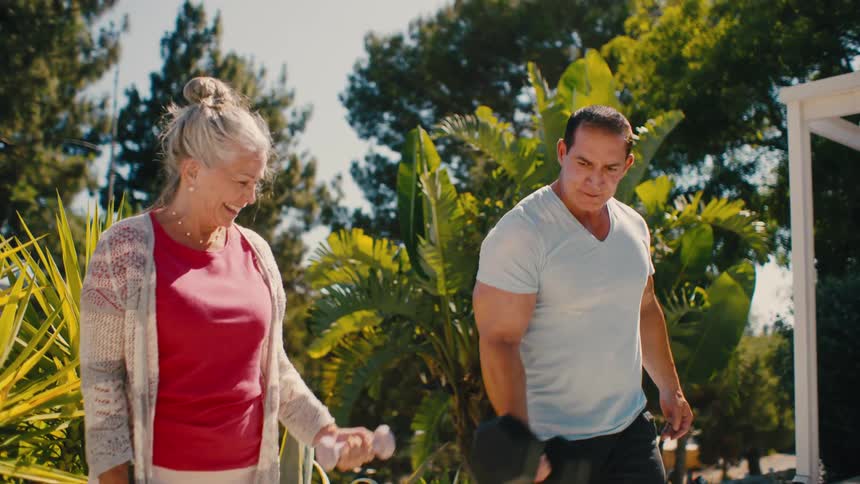 A body builder teaches an elderly woman how to lift weights as she smiles