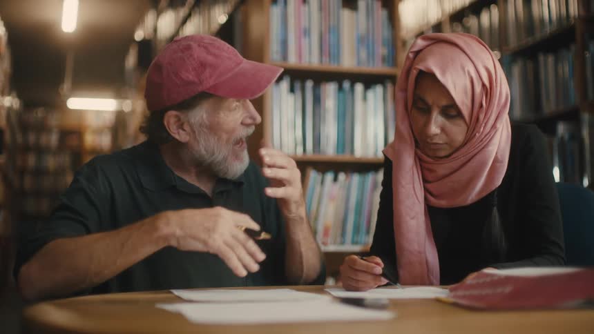 An older man sitting at atable with a younger woman conversing over paperwork