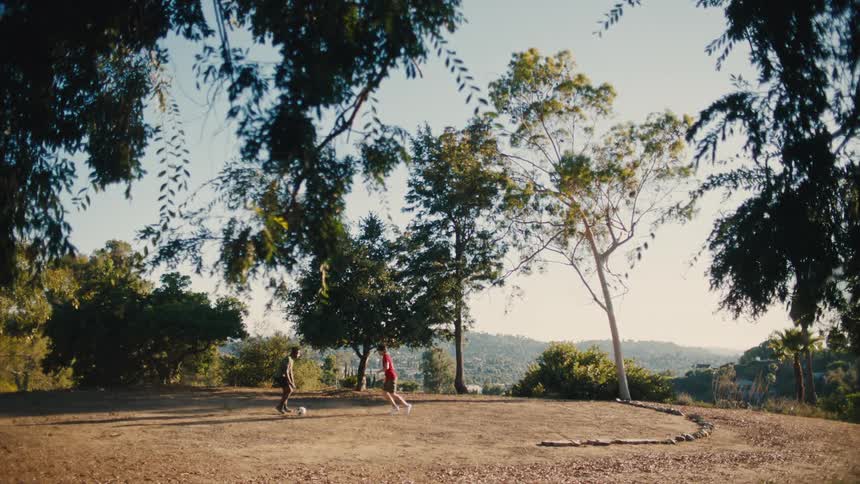 Two boys playing soccer together on a gravel area in the middle of the day