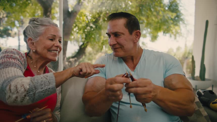 An older woman knitting on a couch next to her adult son who is also trying to knit
