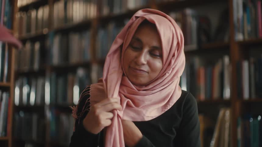 Woman with a scarf around her head holding a pen sitting at a table with a person next to her