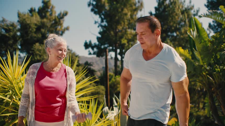 Man and an elderly woman lifting weights with dumbbells in an outdoor setting with trees around