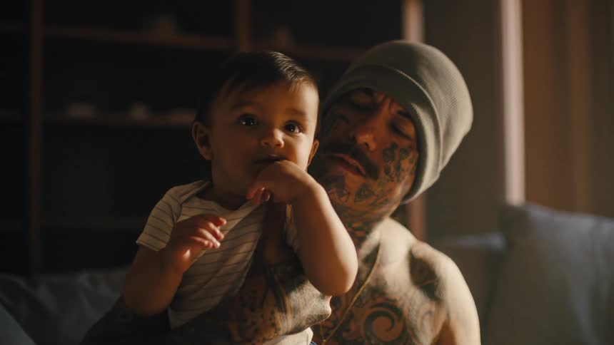 Shirtless tattooed man holds a baby in his arms while seated on a couch in a cozy living room