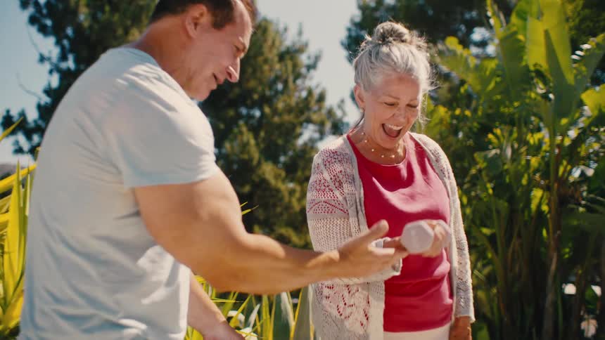 A man fitness trainer helping an older women lift hand weights on a stone patio on a bright sunny day