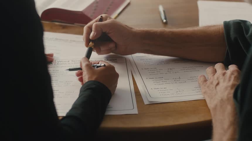 Hands of a man sitting at a table next to a woman whose hands are also on the table as they point to paperwork with their pens