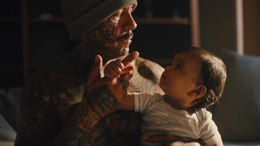 A tattooed man wearing a cap sits on a couch while holding a baby