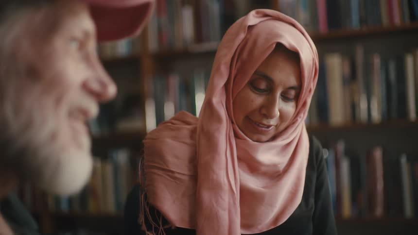 A woman with a pink turban on her head pointing a pen at a paper on a table and there is a man next to her talking to her