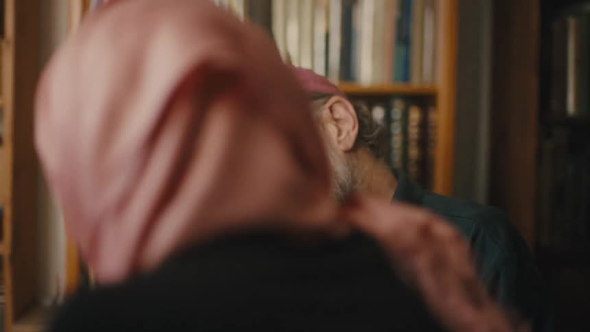 An elderly man in a cap is looking at a paper on a table while talking to a woman standing next to him who is wearing a turban