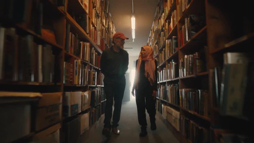 A man wearing a red cap walking with a young woman through a row of books in a library as they conversate