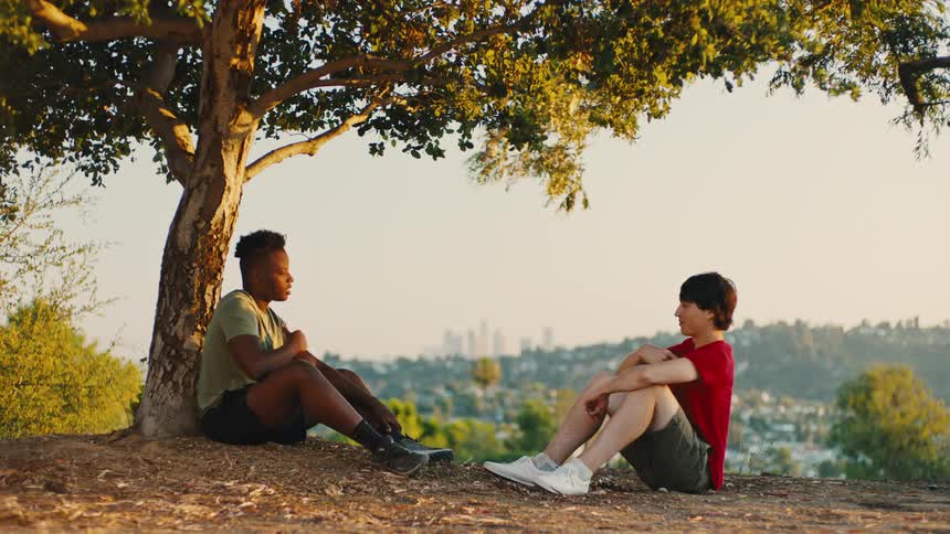 Two friends are sitting under a tree on a grassy hill overlooking a cityscape during the day