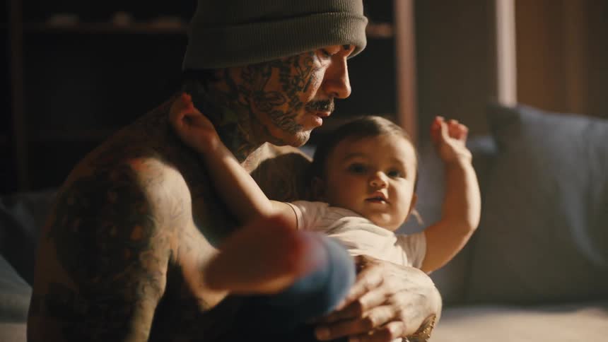 A man wearing a hat and no shirt is holding a little boy and the man has tattoos all over his body