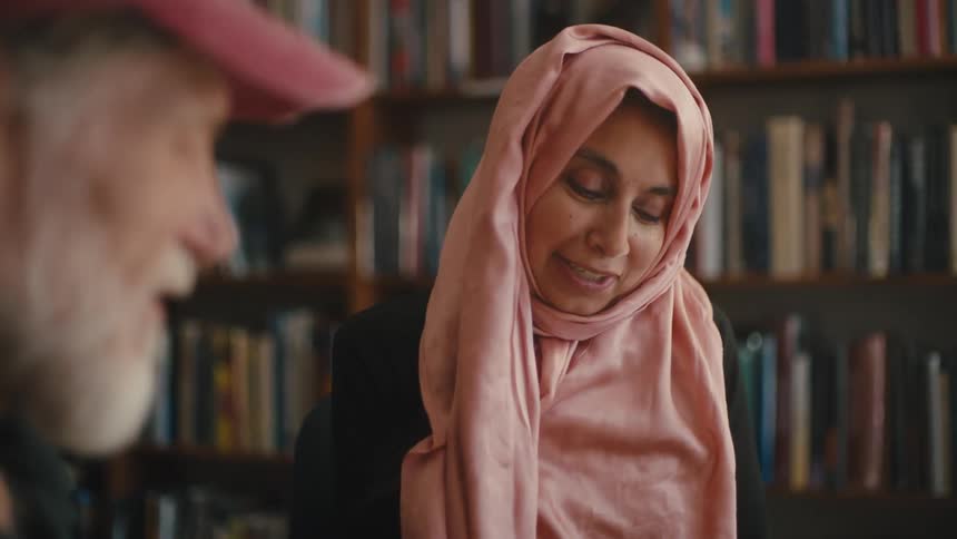 A woman wearing a pink turban is pointing to a paper on a table and a man standing next to her is talking to her and she is smiling