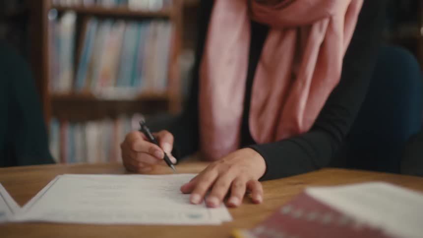 A man in a red cap talking to a woman in a pink scarf while they are sitting at table in a library