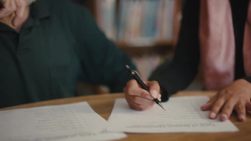 A bearded man explains a document to a woman wearing a pink headscarf in a library