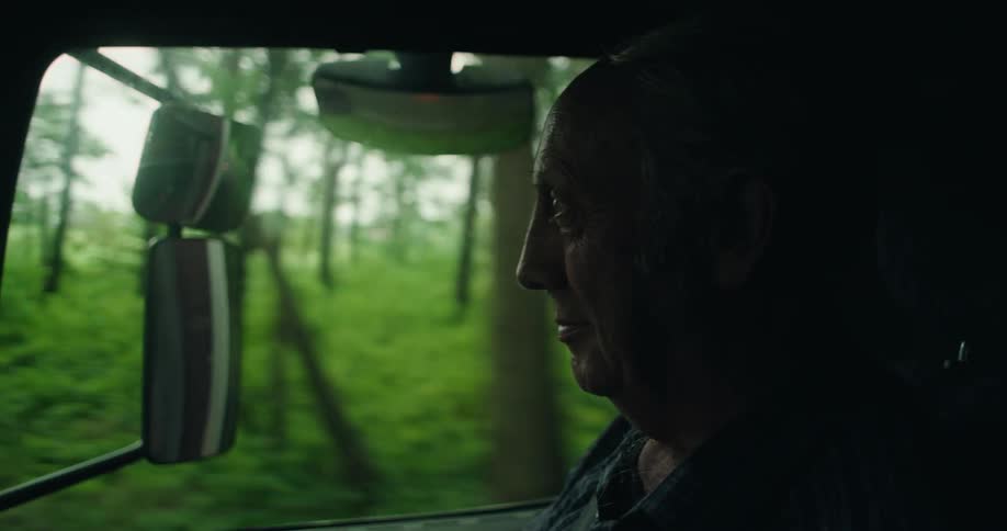 The side profile of an older man driving a vehicle down a road with trees and bushes alongside the road
