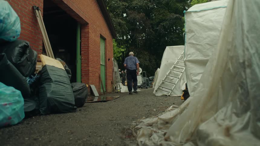 An elderly man walks past a garage door and a pile of garbage bags