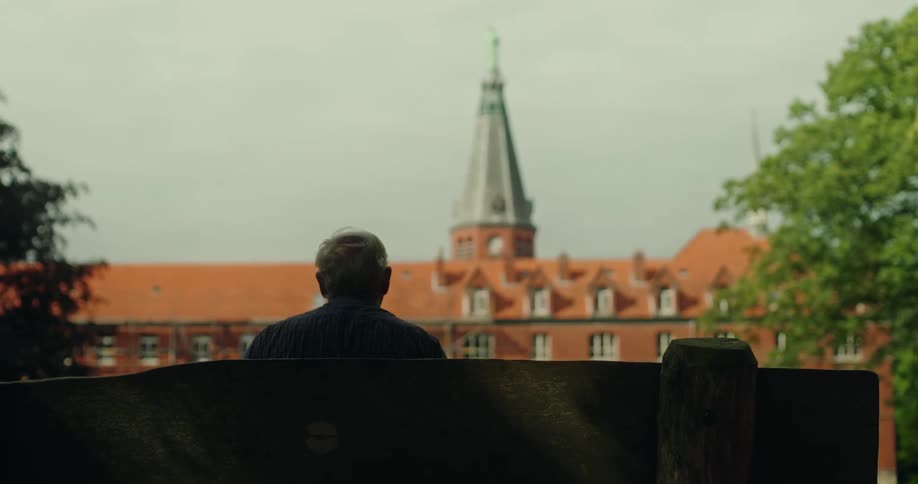 Elderly man sitting on a bench looking at a monastery with a spire in the background