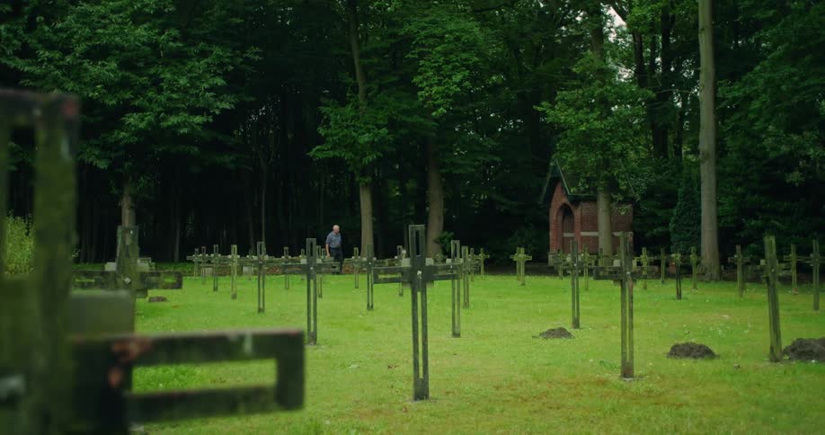 Many crosses displayed in a grassy graveyard surrounded by trees with a man looking over it
