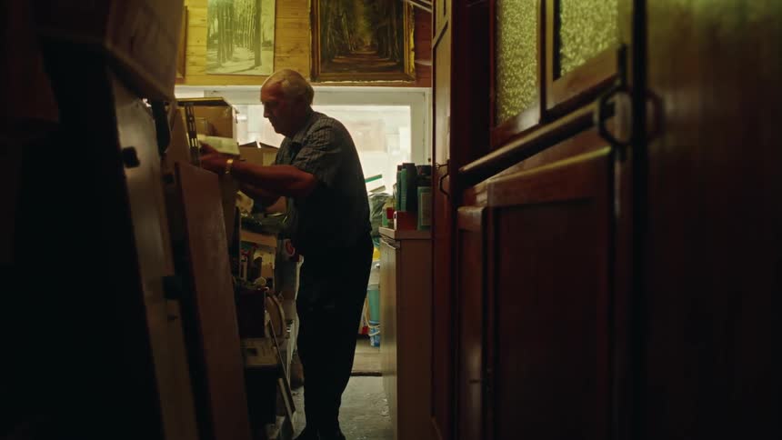 A man carries a cardboard box out of a cluttered room stacked haphazardly
