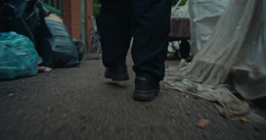 The legs of a person wearing black shoes walking on a floor with various junk on the sides