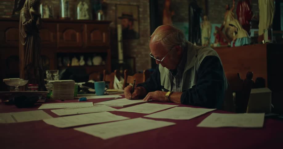 An older man sitting at a table writing on paper with other pieces of paper spread out in front of him