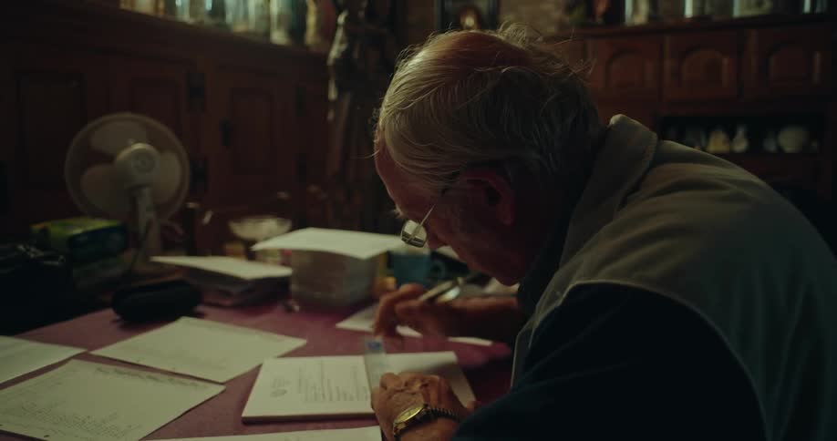 An elderly man sits at a desk in a dimly lit room carefully writing on a document