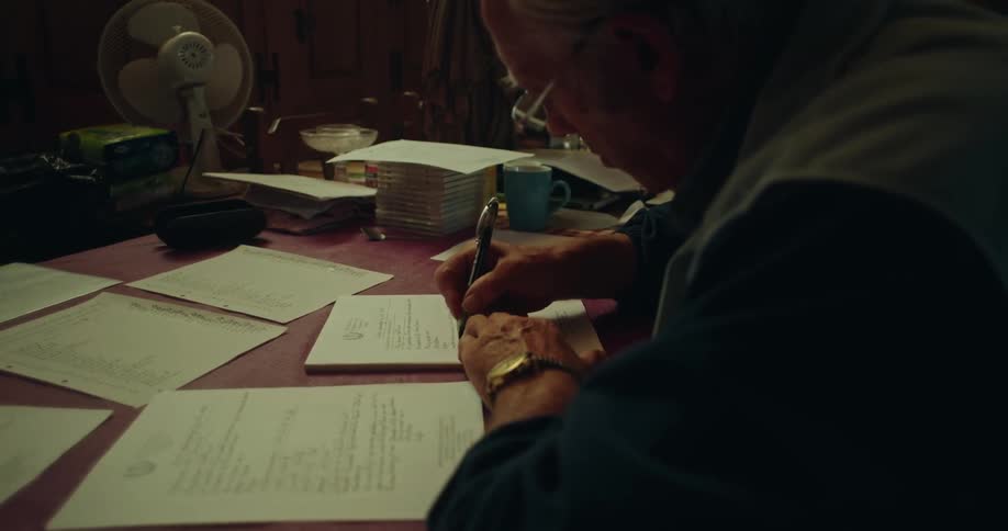 An elderly man wearing glasses is writing on a paper using a pen and there are several papers scattered around the table
