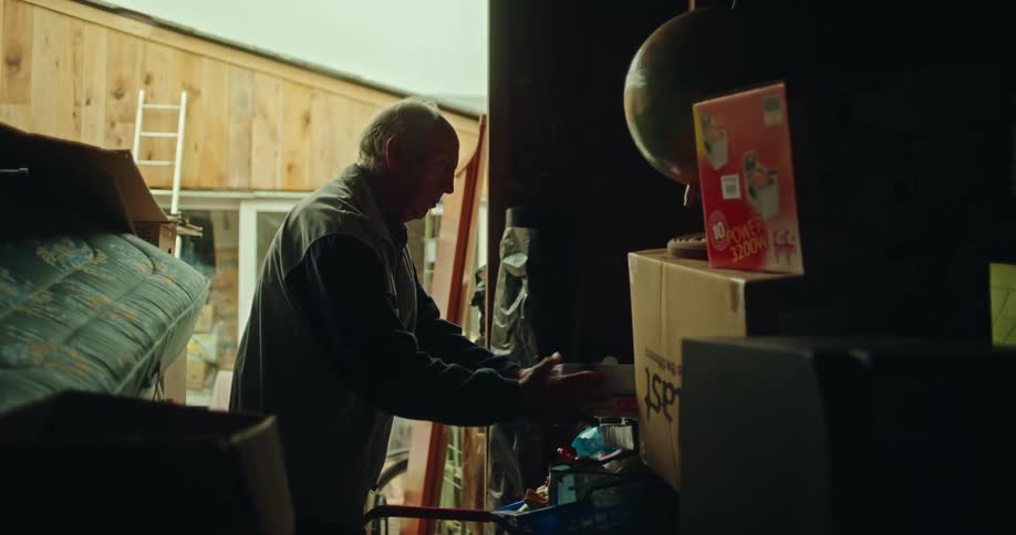 Elderly man storing two boxes in a shed and looking at a scientific globe surrounded by clutter