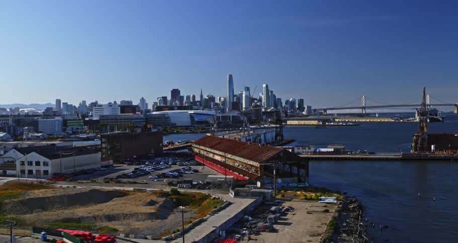 A port with a parking lot with many cars in front of a body of water with a metropolitan city in the background