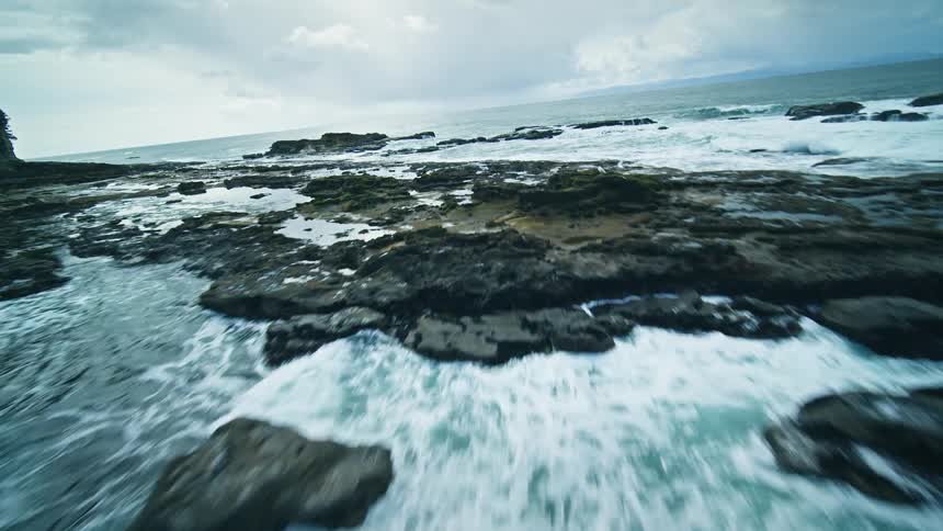 Aerial view of a beach with waves crashing onto the rocky shore and coniferous trees on a cloudy day