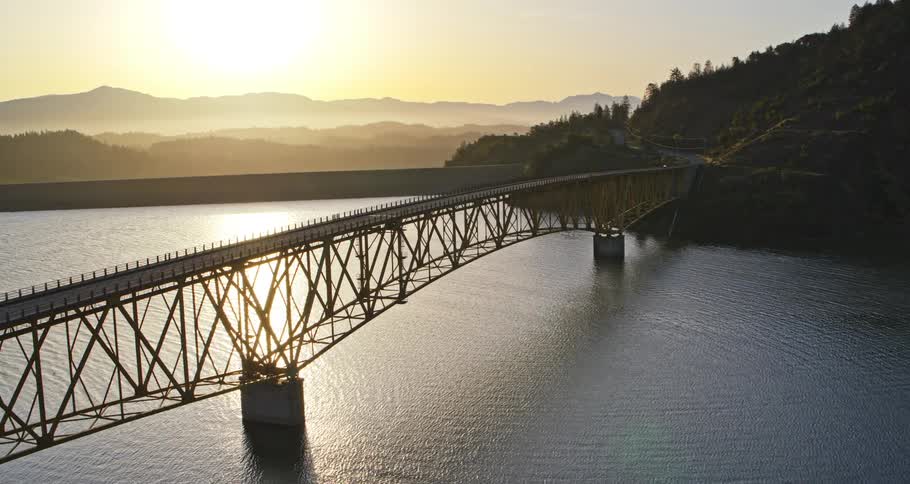 A scenic aerial view shows a bridge spanning a lake at sunset with mountains in the background