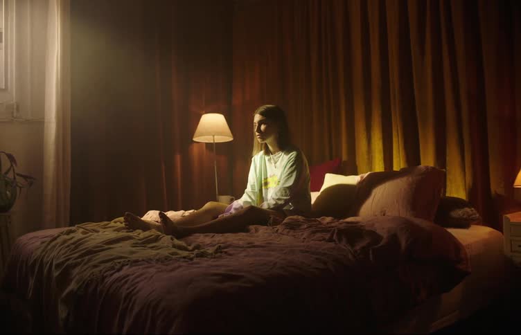 A woman sits in a rumpled bed with her face turned to the sunlight streaming in through a window