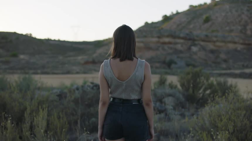 Woman walks through a field towards the water at the base of the hills