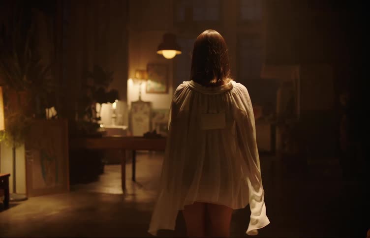 A woman in a white dress is walking into a living room that is lit with some yellow lights and she stops