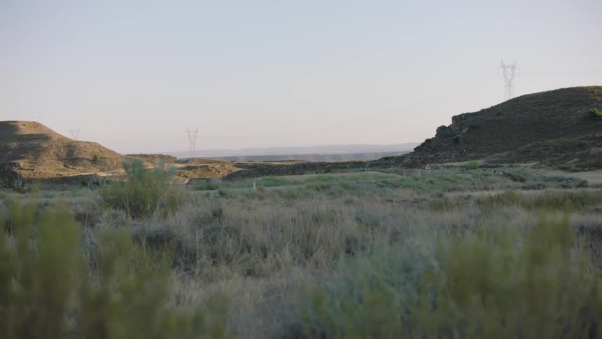 A wide shot shows a dry grassy landscape with hills in the background and power lines crossing the horizon