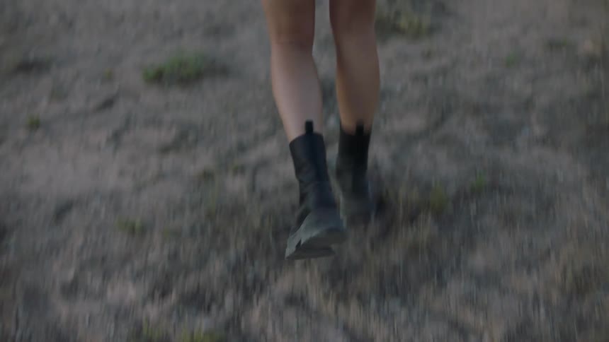 Woman in skirt and boots walking through a field with mountains in the background